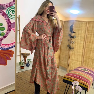 Puede incluir: Una bata larga y fluida con un estampado floral en tonos verdes y rosas. La bata tiene mangas anchas y un cinturón a juego. El modelo lleva botas azul oscuro. La bata mide aproximadamente 140 cm de largo.