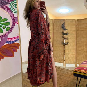 May include: A long, flowing red kimono-style robe with a paisley pattern in black and other colors. The robe is open in the front and has long sleeves. The person in the image is wearing high-heeled shoes.