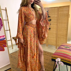 Puede incluir: Conjunto de dos piezas con estampado floral naranja. El top es un crop top con escote pronunciado y mangas largas con volantes. La falda tiene una abertura alta y detalles de borlas. Estilo bohemio.