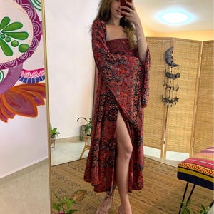 May include: A long, red dress with a square neckline and flared sleeves. The dress features a paisley pattern in black and red. The dress has a high slit on the left side. The person is wearing clear heels.