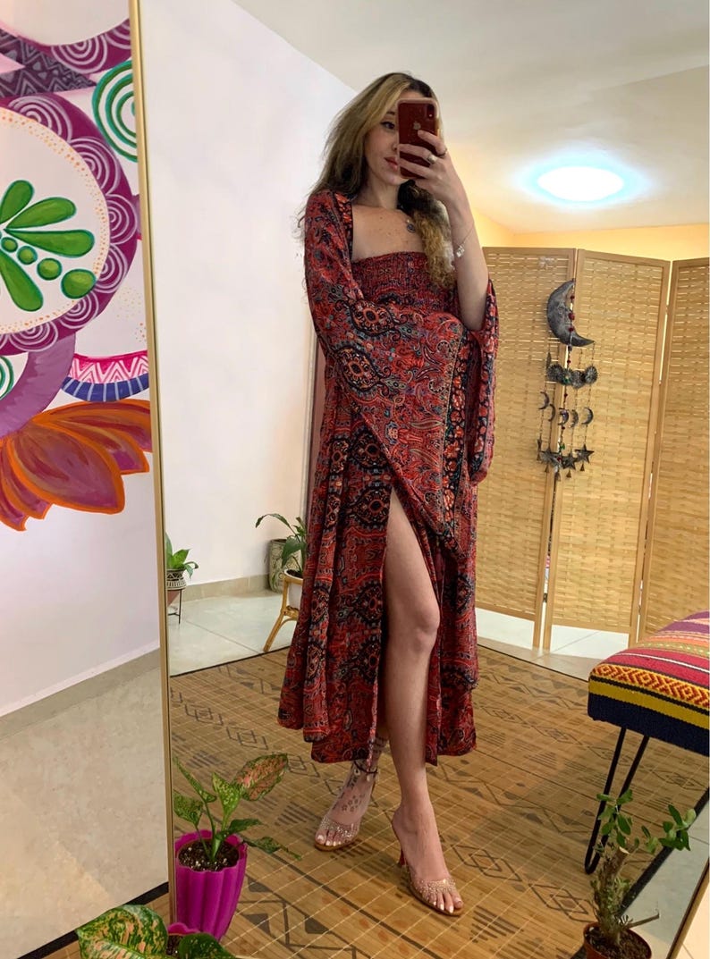 May include: A long, red dress with a square neckline and flared sleeves. The dress features a paisley pattern in black and red. The dress has a high slit on the left side. The person is wearing clear heels.