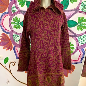 May include: A hooded tunic dress in a deep burgundy color with a paisley pattern in gold. The dress has long sleeves and a hood with a collar. The hemline and cuffs feature a decorative border. The dress is displayed on a mannequin.