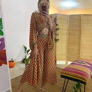 May include: A red and gold patterned two-piece outfit with a crop top and wide-leg pants. The top has a knotted detail at the front and the pants have a wide leg and a split hem.