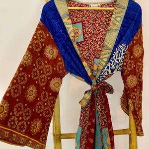 May include: A colorful kimono-style robe with long sleeves, hanging on a wooden hanger. The robe features a mix of patterns and colors, including burgundy, blue, and gold. The sleeves and front panels have intricate floral and geometric designs.