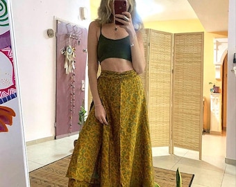 May include: A woman wearing a green tank top and a long, yellow, floral print wrap skirt. The skirt has multiple layers and a flowing silhouette. She is wearing brown boots and has her hair down.