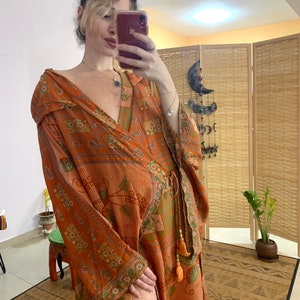 Pode incluir: Robe laranja de mangas compridas com um padrão floral e geométrico. O robe tem decote em V e cintura com cordão com borlas laranja. As mangas e as bordas são adornadas com uma borda verde e branca. Feito de tecido leve.