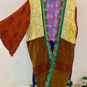 Puede incluir: Una colorida bata estilo kimono con patchwork. La bata presenta una mezcla de patrones y colores, incluyendo mangas rojas con estampado de cachemira, flores doradas, rayas marrones y telas florales azules. La bata está colgada en una percha de madera.