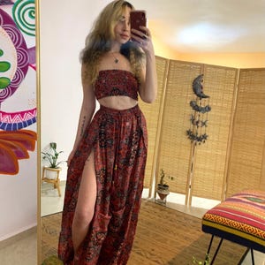 May include: A woman wearing a red and black patterned crop top and matching pants with a side slit. The pants have a drawstring waist and a loose, flowy fit. The woman is standing in front of a mirror and a wooden screen.