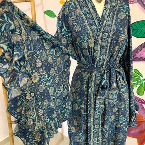 May include: A long, flowing robe in shades of navy blue, teal, and tan, adorned with a floral pattern. The robe features wide sleeves with a ruffled edge and a matching belt. The garment is displayed on a mannequin, showcasing its elegant design.