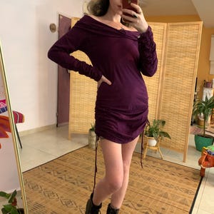 May include: A dark purple, off-the-shoulder dress with long sleeves and a ruched, drawstring design. The dress is paired with black combat boots. The background includes a mirror, a folding screen, and potted plants.