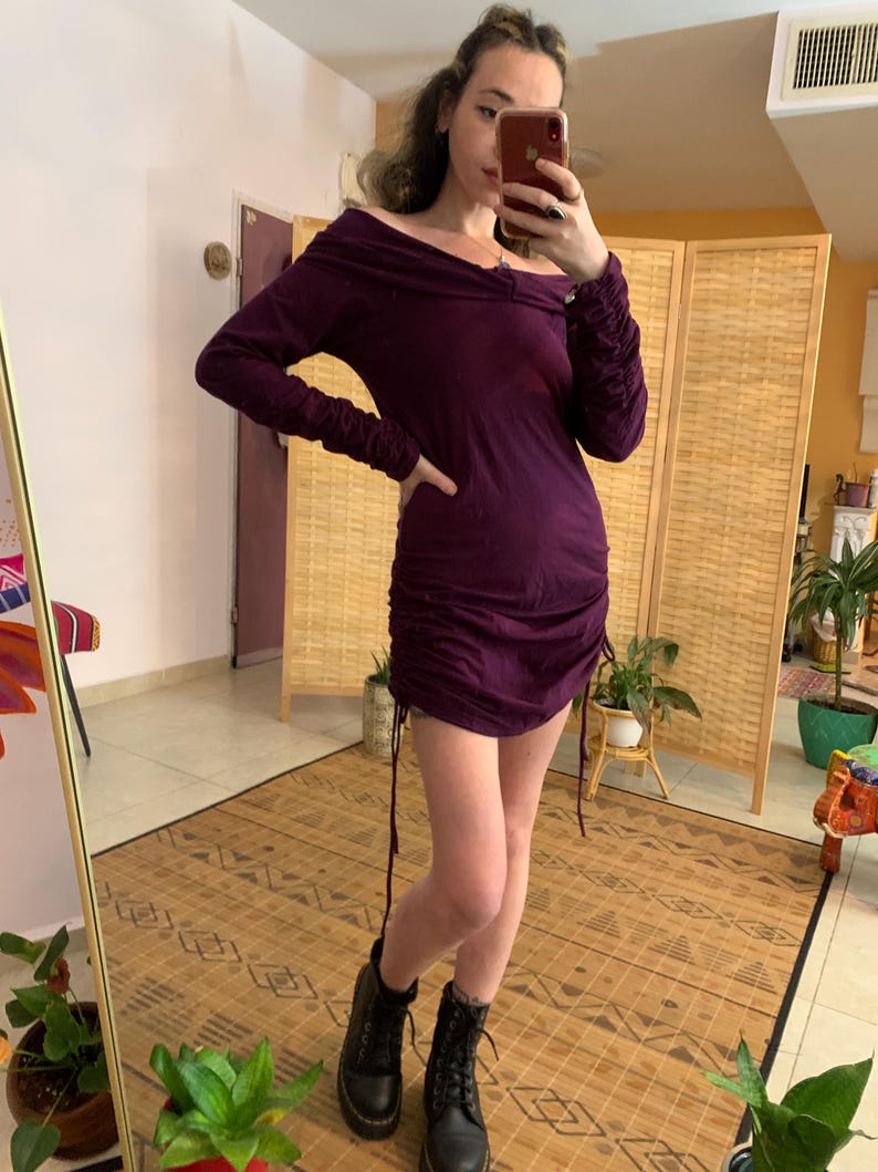 May include: A dark purple, off-the-shoulder dress with long sleeves and a ruched, drawstring design. The dress is paired with black combat boots. The background includes a mirror, a folding screen, and potted plants.