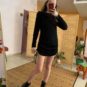 May include: A black long-sleeve dress with a cowl neck and drawstring details on the sides. The dress is paired with black combat boots. The image is taken in front of a mirror.