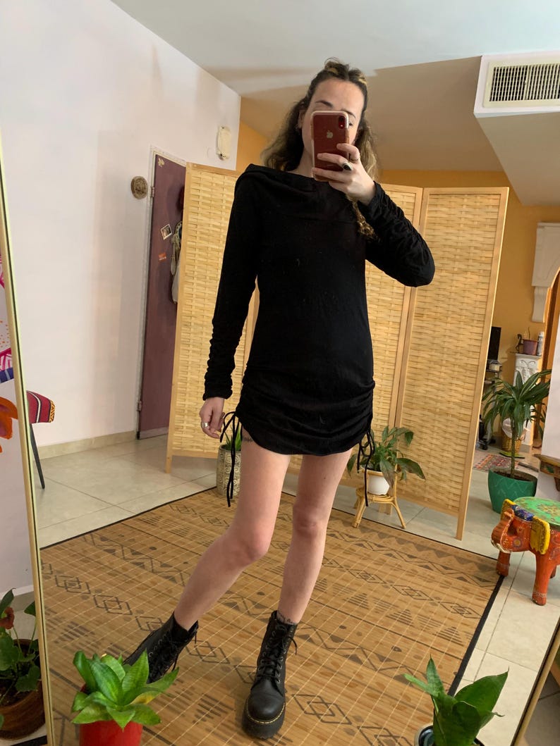 May include: A black long-sleeve dress with a cowl neck and drawstring details on the sides. The dress is paired with black combat boots. The image is taken in front of a mirror.