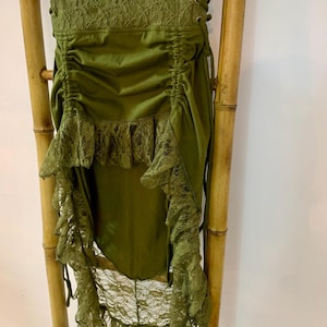 May include: Olive green high-low skirt with lace trim and a ruched waist. The skirt features a lace-up detail on the side and a layered, asymmetrical hemline. The skirt is hanging on a bamboo ladder.