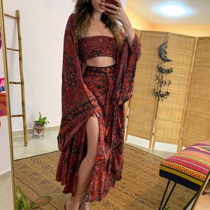 May include: A red and black patterned two-piece set, including a crop top and a long skirt with a high slit. The set features a paisley-like design. The sleeves are long and flowing. The outfit is paired with clear sandals.
