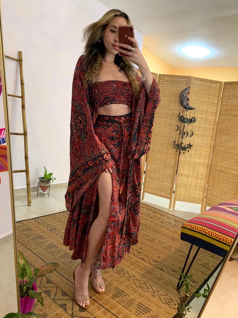 May include: A red and black patterned two-piece set, including a crop top and a long skirt with a high slit. The set features a paisley-like design. The sleeves are long and flowing. The outfit is paired with clear sandals.