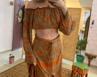 Könnte beinhalten: Ein zweiteiliges Outfit, bestehend aus einem schulterfreien Crop-Top und einem passenden Rock. Das Set zeigt ein Blumenmuster in Orange-, Braun- und Goldtönen. Der Rock hat einen Wickel-Design. Das Ensemble ist aus leichtem Stoff gefertigt.