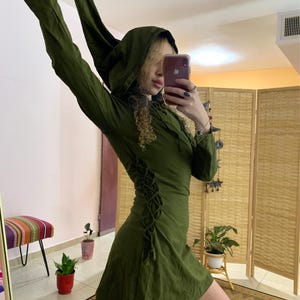 以下が含まれることがあります： サイドにレースアップのディテールが付いた緑色のフード付きドレス。ドレスは短く、長袖です。