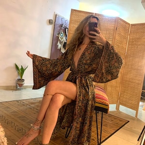 May include: A long, black and gold floral print robe with a deep V-neckline and long sleeves. The robe is tied at the waist with a matching sash. The model is wearing the robe and sitting on a stool.