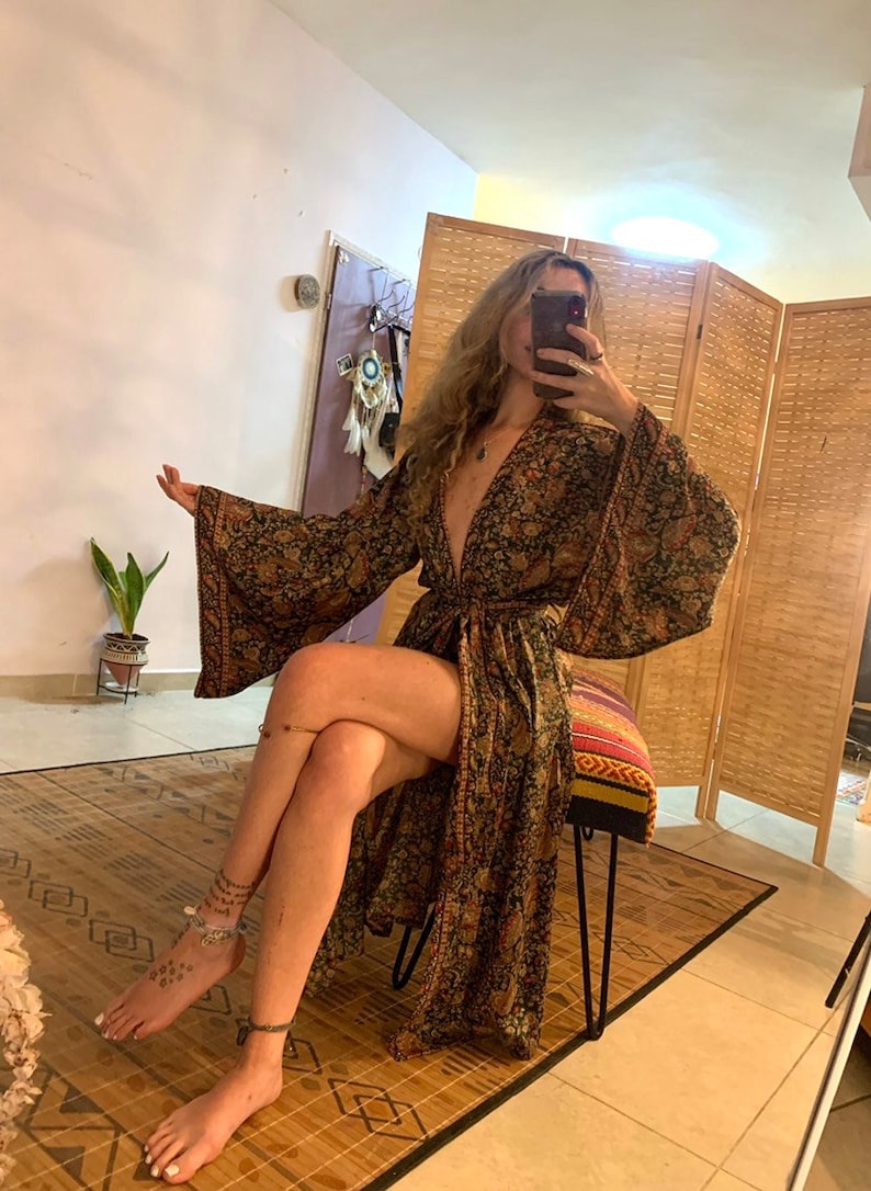 May include: A long, black and gold floral print robe with a deep V-neckline and long sleeves. The robe is tied at the waist with a matching sash. The model is wearing the robe and sitting on a stool.