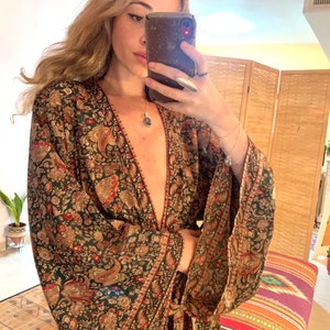 Könnte beinhalten: Eine lange, fließende Robe in Grün und Gold mit Paisley-Muster, tiefem V-Ausschnitt und langen Ärmeln. Die Robe wird mit einem Band in der Taille gebunden.