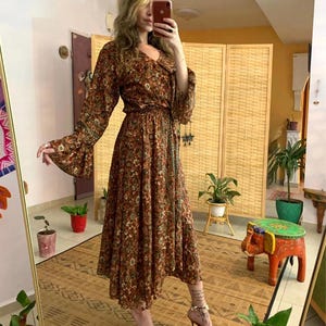 May include: A long-sleeved maxi dress with a floral print in shades of brown, green, and orange. The dress has a V-neckline, a fitted bodice, and a flowing skirt. The dress is cinched at the waist with a tie.