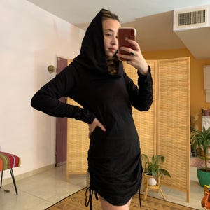 May include: Black hooded dress with long sleeves and a ruched, drawstring hem. The dress is made of a soft, dark fabric and has a fitted silhouette. The model is holding a phone and is standing in front of a woven room divider.
