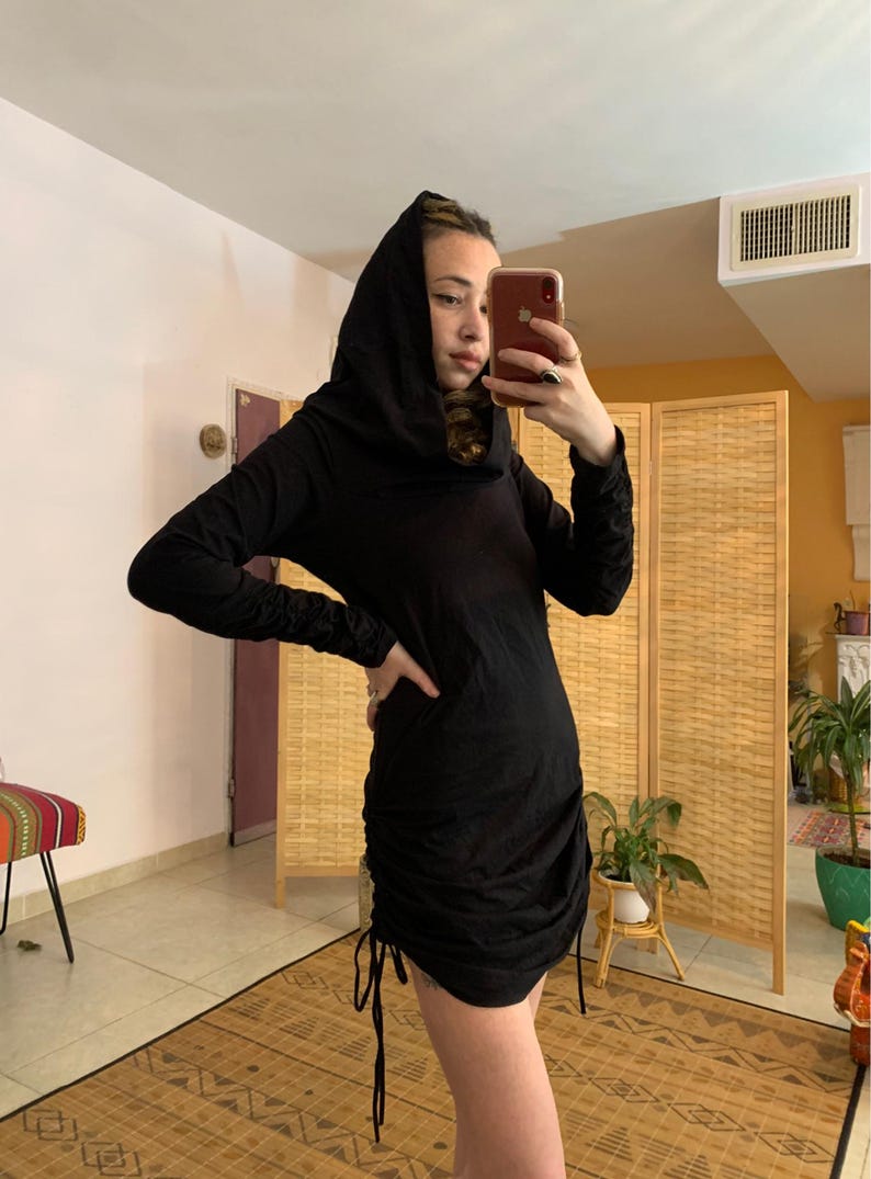 May include: Black hooded dress with long sleeves and a ruched, drawstring hem. The dress is made of a soft, dark fabric and has a fitted silhouette. The model is holding a phone and is standing in front of a woven room divider.