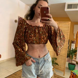 Op de afbeelding: Een bruin en groen bloemenprint off-shoulder crop top met lange bell sleeves. De top is gemaakt van een lichtgewicht stof en heeft een ontspannen pasvorm.