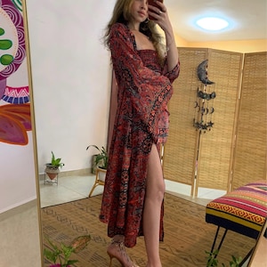 May include: A long, red dress with a paisley pattern and a high slit. The dress has long, flared sleeves and a square neckline. The person is wearing clear heels. The dress is a bohemian style.