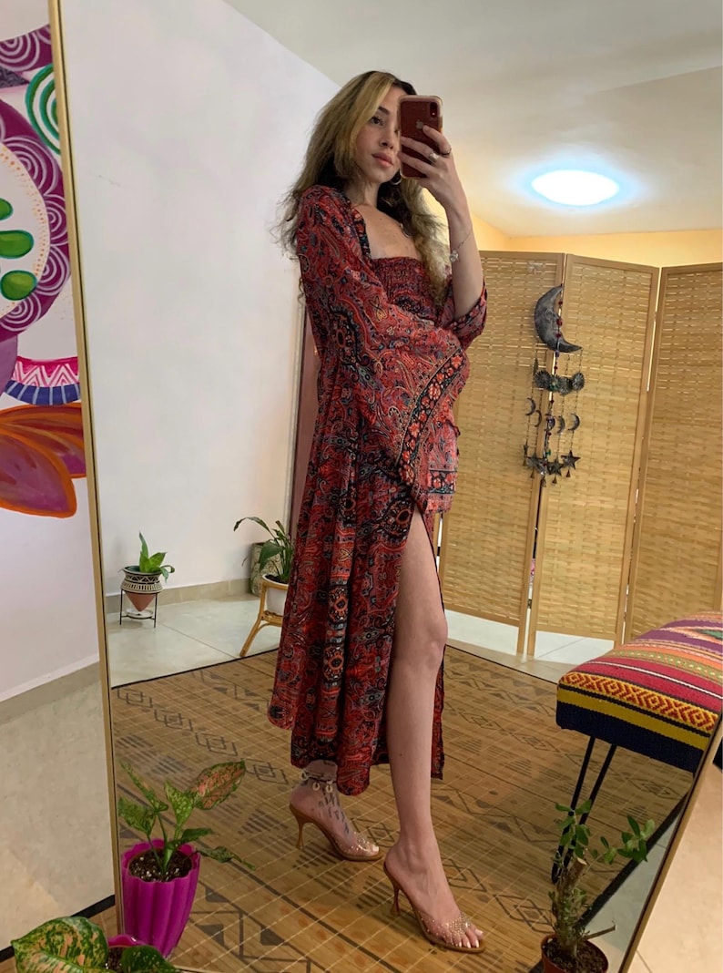 May include: A long, red dress with a paisley pattern and a high slit. The dress has long, flared sleeves and a square neckline. The person is wearing clear heels. The dress is a bohemian style.