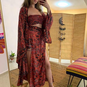 May include: A three-piece ensemble featuring a red and black paisley print. The set includes a strapless crop top, a long-sleeved kimono, and a wrap skirt with a high slit. The outfit is worn with high-heeled sandals.