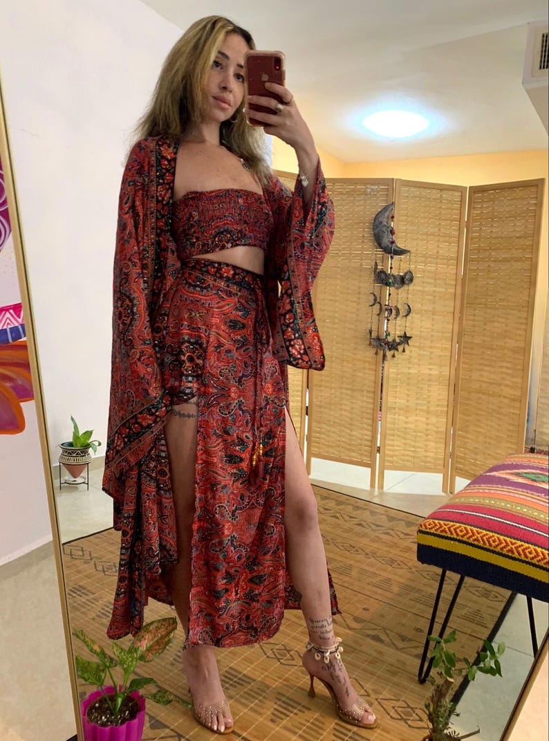 May include: A three-piece ensemble featuring a red and black paisley print. The set includes a strapless crop top, a long-sleeved kimono, and a wrap skirt with a high slit. The outfit is worn with high-heeled sandals.