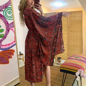 Könnte beinhalten: Ein langes, fließendes rotes Kleid mit Paisley-Muster und schwarzen Akzenten. Das Kleid hat weite Ärmel und ein Wickeldesign. Das Kleid wird mit hochhackigen Sandalen getragen. Das Gesamtthema ist Bohème-Mode.