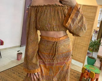 Könnte beinhalten: Ein zweiteiliges Outfit, bestehend aus einem schulterfreien Crop-Top und einem Wickelrock. Das Set zeigt ein Paisley-Muster in Gold-, Orange- und Rosatönen. Das Oberteil hat lange, ausgestellte Ärmel. Der Rock ist im Wickelstil mit Bindeband.