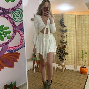 May include: A white, long-sleeved crop top with a tie front and a white, asymmetrical skirt with a lace trim. The skirt has a tie detail on the side. The model is wearing green ankle boots.