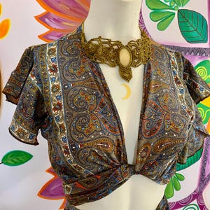 May include: A paisley-patterned crop top and matching skirt in shades of brown, blue, orange, and white are displayed on a mannequin. A woven necklace with a central stone and a crescent moon pendant is worn with the outfit. The background features colorful floral artwork.