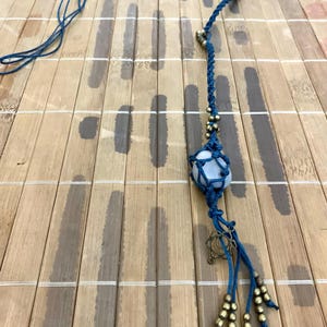 May include: A handcrafted macramé pendant necklace with a light blue stone in a woven blue cord. The necklace has gold-coloured beads and a decorative charm, with a long cord. The pendant is displayed on a bamboo surface.