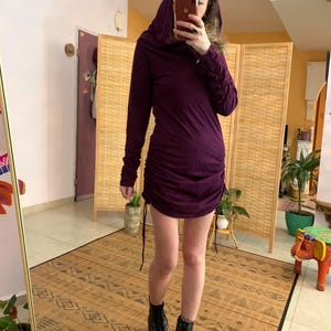 May include: A dark purple hooded dress with long sleeves and a ruched design. The dress is paired with black combat boots. The person is standing in front of a mirror and a folding screen.