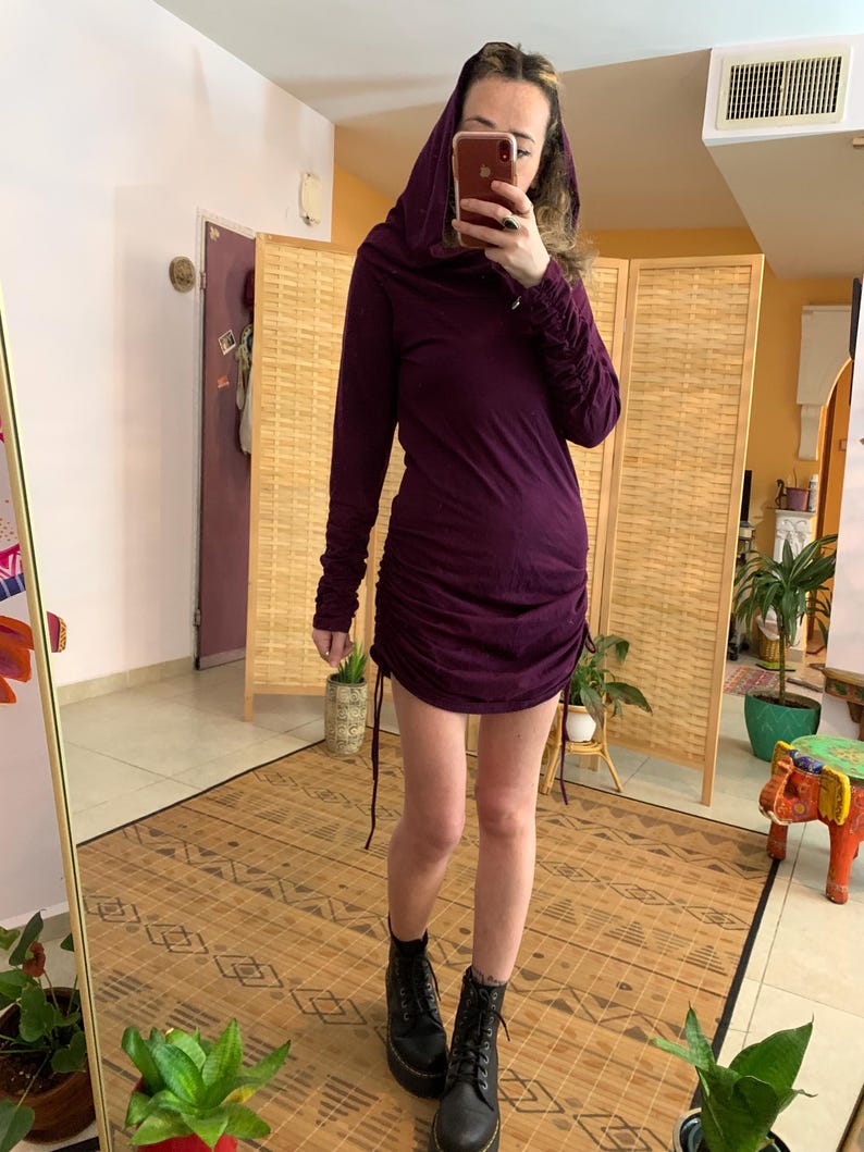 May include: A dark purple hooded dress with long sleeves and a ruched design. The dress is paired with black combat boots. The person is standing in front of a mirror and a folding screen.