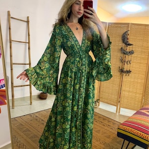 Puede incluir: Un vestido largo y fluido de color verde con un estampado floral. El vestido presenta mangas anchas y acampanadas y un escote en V profundo. La tela parece ligera y el vestido está recogido en la cintura. El vestido es largo hasta el suelo.