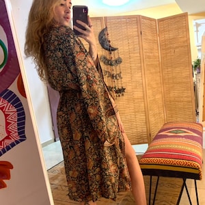 May include: A woman wearing a long, black and brown patterned robe with a belt. The robe has a floral design and a side slit. The woman is standing in front of a mirror and a wooden screen.