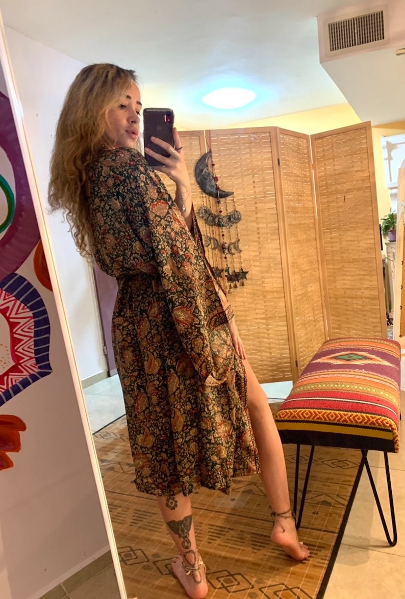 May include: A woman wearing a long, black and brown patterned robe with a belt. The robe has a floral design and a side slit. The woman is standing in front of a mirror and a wooden screen.