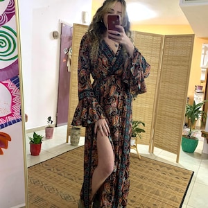 May include: A long, black maxi dress with a paisley print and a side slit. The dress has a V-neckline and long sleeves with bell cuffs. The model is wearing the dress with brown ankle boots.