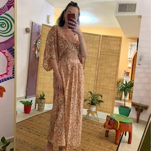 May include: A woman wearing a long, flowing, peach-colored dress with a paisley pattern. The dress has a cut-out detail at the waist and a V-neckline. The dress is made of a lightweight fabric and has a relaxed fit.