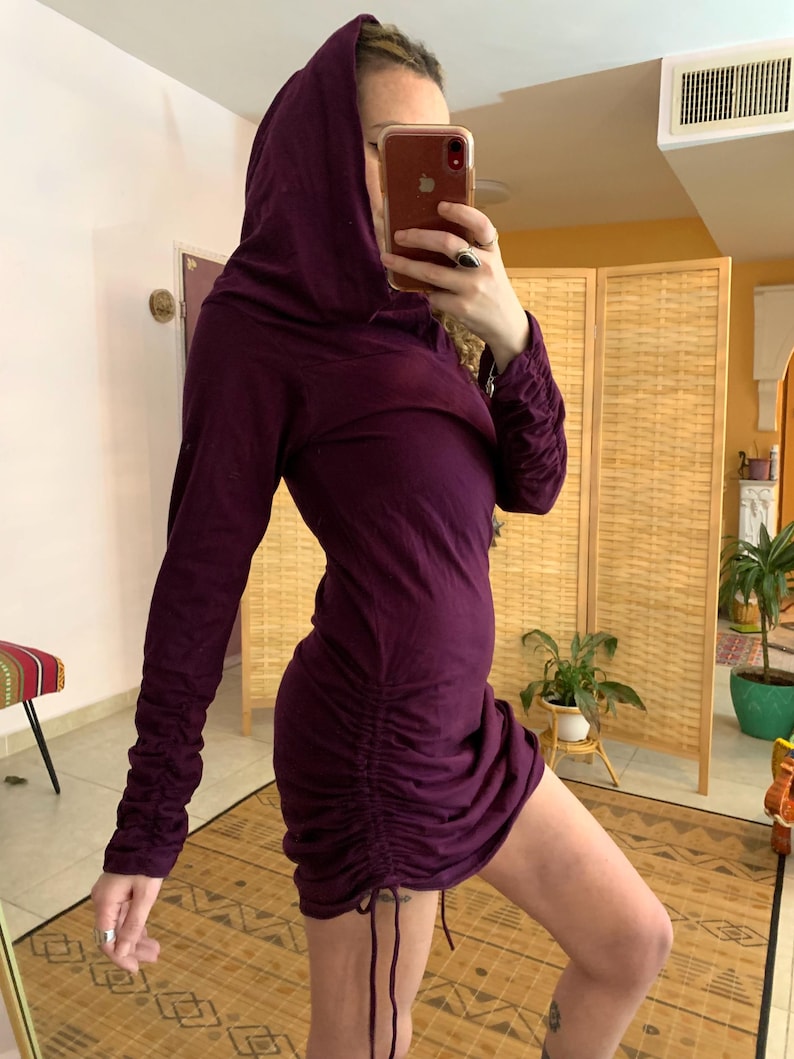 May include: A long-sleeved, hooded dress in a deep purple hue. The dress features a ruched design on the side, with drawstring details. The sleeves are long and gathered at the wrists. The dress is a mini length.