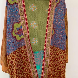 Puede incluir: Una bata tipo kimono patchwork con una variedad de patrones y colores. La bata presenta una mezcla de diseños florales y geométricos en tonos marrón, azul, verde y rojo. La bata está colgada en una percha de madera.