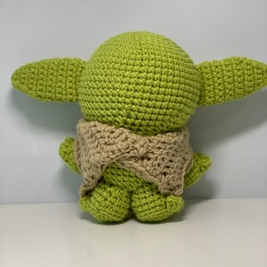 May include: A green crocheted plush toy with a brown crocheted scarf. The toy is shaped like a character with large ears and a round head.
