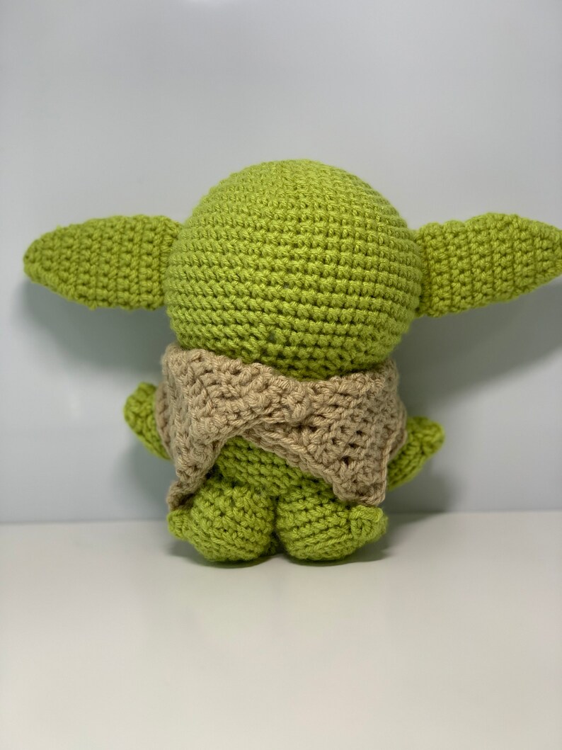 May include: A green crocheted plush toy with a brown crocheted scarf. The toy is shaped like a character with large ears and a round head.