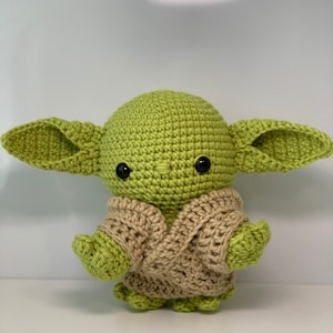 May include: A green crocheted plush toy with large ears and a brown crocheted vest. The toy has black eyes and a small mouth.